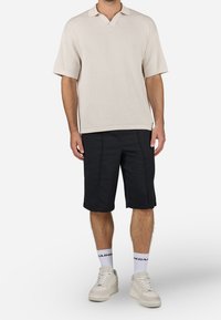 Hombre vestido con una camiseta de punto de manga corta beige, pantalones cortos negros hasta la rodilla, calcetines blancos de marca y zapatillas beige, de pie frente a un fondo liso.