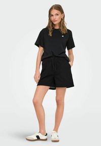 Schwarzes Baumwoll-T-Shirt und schwarze Shorts mit einem lockeren Schnitt und einem Kordelzugbund, kombiniert mit schwarz-weißen Sneakers.
