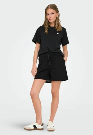 Sort bomulds t-shirt og sorte shorts med løs pasform og snor i taljen, parret med hvide og sorte sneakers.
