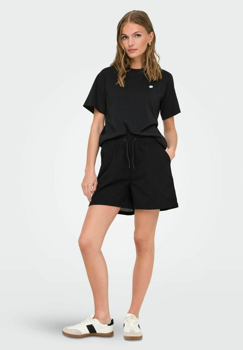 Schwarzes Baumwoll-T-Shirt und schwarze Shorts mit einem lockeren Schnitt und einem Kordelzugbund, kombiniert mit schwarz-weißen Sneakers.