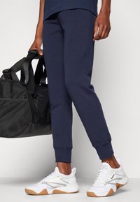 Person, die marineblaue Jogginghosen und weiße Reebok-Sneaker trägt, hält eine schwarze Sporttasche an ihrer Seite vor einem schlichten Hintergrund.