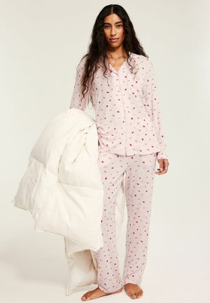 Vrouw met lang, krullend haar staat op blote voeten, draagt lichtroze pyjama met patroon, en houdt een grote witte gewatteerde deken over één arm.