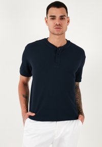 Tengerészkék, rövid ujjú henley póló, texturált anyagból, kerek gallérral és három gombos lezárással, fehér nadrággal viselve.