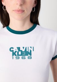 White T-shirt with turquoise ribbed neckline and cuffs, featuring a prominent embroidered graphic "CALVIN KLEIN 1968" in matching color.