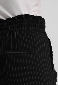 Pantalons noirs à rayures avec une taille froncée et des détails de poches latérales. Le tissu semble lisse, structuré et léger.