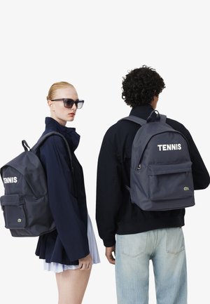 Due persone che indossano giacche scure e zaini blu navy con l'etichetta "TENNIS", una rivolta verso l'obiettivo e l'altra di spalle, su sfondo bianco.
