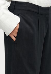 Pantalon sombre et ajusté en tissu lisse. Comprend des poches latérales, une coupe plate à l'avant et une coupe slim, porté avec une chemise blanche boutonnée.