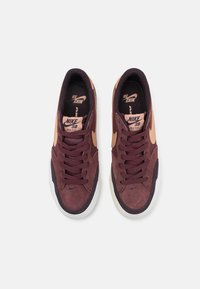 Adidași din suede maroniu cu talpă albă. Prezintă șireturi și un logo Nike de culoare roz deschis. Partea superioară texturată cu branding pe limba încălțămintei.