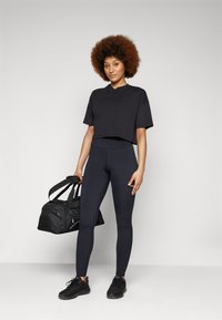 T-shirt nera corta, leggings blu scuro a vita alta, sneakers nere, che tiene in mano una borsa da palestra nera. Texture liscia, design sportivo e casual.