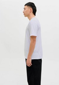 Valkoinen lyhythihainen T-paita pyöreällä pääntiellä, valmistettu puuvillasta, esitetty henkilöllä seisomassa kyljelleen, yhdistettynä mustiin housuihin.