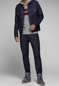 Chaqueta de mezclilla en azul oscuro con dos bolsillos frontales, superpuesta a una sudadera gris con un logo burdeos, combinada con unos pantalones de mezclilla oscuros y botas grises.