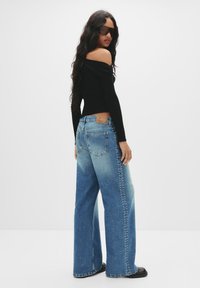 Zwarte off-shoulder top gecombineerd met blauwe wijde jeans, met een vervaagde afwerking en studaccenten langs de zijnaad. Zwarte schoenen.