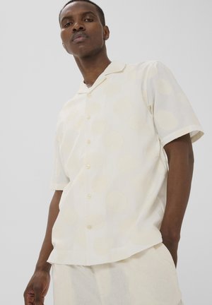 Mand iført kortærmet cremefarvet skjorte med subtile cirkelmønstre og lyse bukser, stående foran en ensfarvet baggrund.