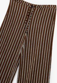 Bruine broek met verticale witte en zwarte strepen, voorzien van een verstelbare tailleband op een witte achtergrond.