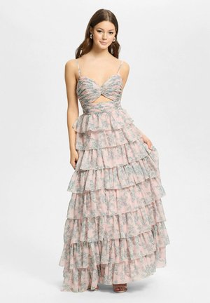 Jonge vrouw die een roze maxi-jurk met bloemmotief en meerdere lagen draagt, met dunne bandjes en een uitsnijding aan de voorkant, staand tegen een effen lichte achtergrond.