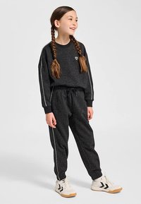 Czarny komplet bluzy i spodni w białe kropki z białymi paskami, z małym logo w kształcie serca. Modelka ma na sobie białe sneakersy z czarnymi detalami.