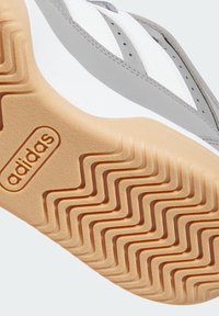 Biele a šedé tenisky s textúrovanou gumovou podrážkou, ktorá má vlnitý vzor a embosované logo "adidas". Hladký syntetický zvršok a šnúrky.