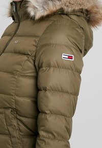 Chaqueta acolchada color verde oliva con capucha con borde de piel, textura acolchada y un parche con logo en la manga que presenta rayas azules, blancas y rojas.