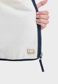 Fleece-Jacke innen, weißer Stoff, mit dunkelblauem Reißverschluss und Besatz. Verfügt über ein T-KIT-Label in Beige mit braunem Text. Sanfte Textur.
