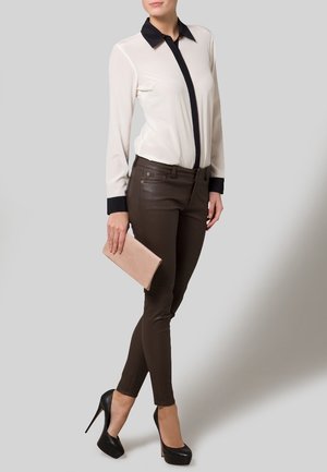 Frau trägt eine weiße Bluse mit schwarzem Kragen und Manschetten, braune eng anliegende Hose, schwarze High Heels und hält eine beige Clutch-Handtasche.