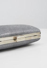 Pochette en tissu métallique argenté avec fermoir doré gravé du mot "MASCARA" sur un fond clair uni.