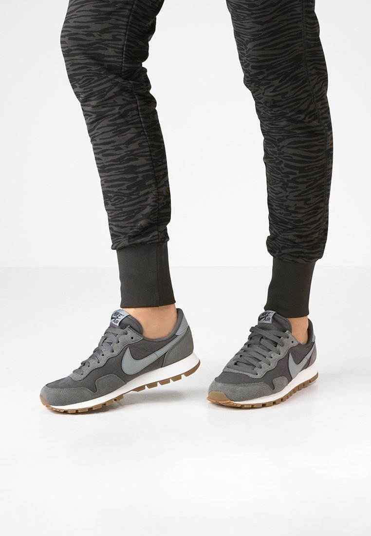 Sneaker Nike grigie realizzate in materiali scamosciato e mesh, con una suola intermedia bianca, logo swoosh grigio e una suola testurizzata. Abbinati a pantaloni jogger con fantasia.