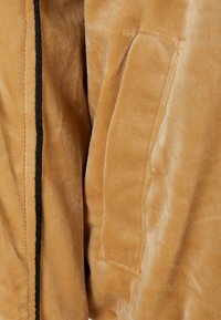 Gros plan d'une veste en daim beige montrant une texture lisse, une couture, une poche et un détail de bordure noire le long du bord.