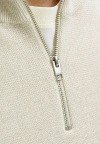 Svijetlosmeđi zip-up pulover s teksturiranim pletenim dizajnom i visokim ovratnikom. Ima srebrni patentni zatvarač s detaljem brendiranja. Nosi se preko kremaste majice.