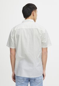 Camisa de hombre de manga corta en tejido beige claro; diseño casual con canesú en la parte posterior y cuello doblado, combinada con jeans de mezclilla azules.