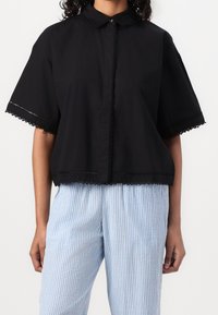 Camicia nera di cotone con maniche corte, finitura in pizzo sui bordi, chiusura con bottoni, design corto; abbinata a pantaloni a righe blu chiaro e bianchi.