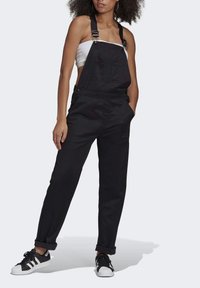 Černé denimové lacláče s nastavovacími popruhy, přední kapsou a volným střihem; doplněné černobílými teniskami se špičkou ve tvaru mušla.