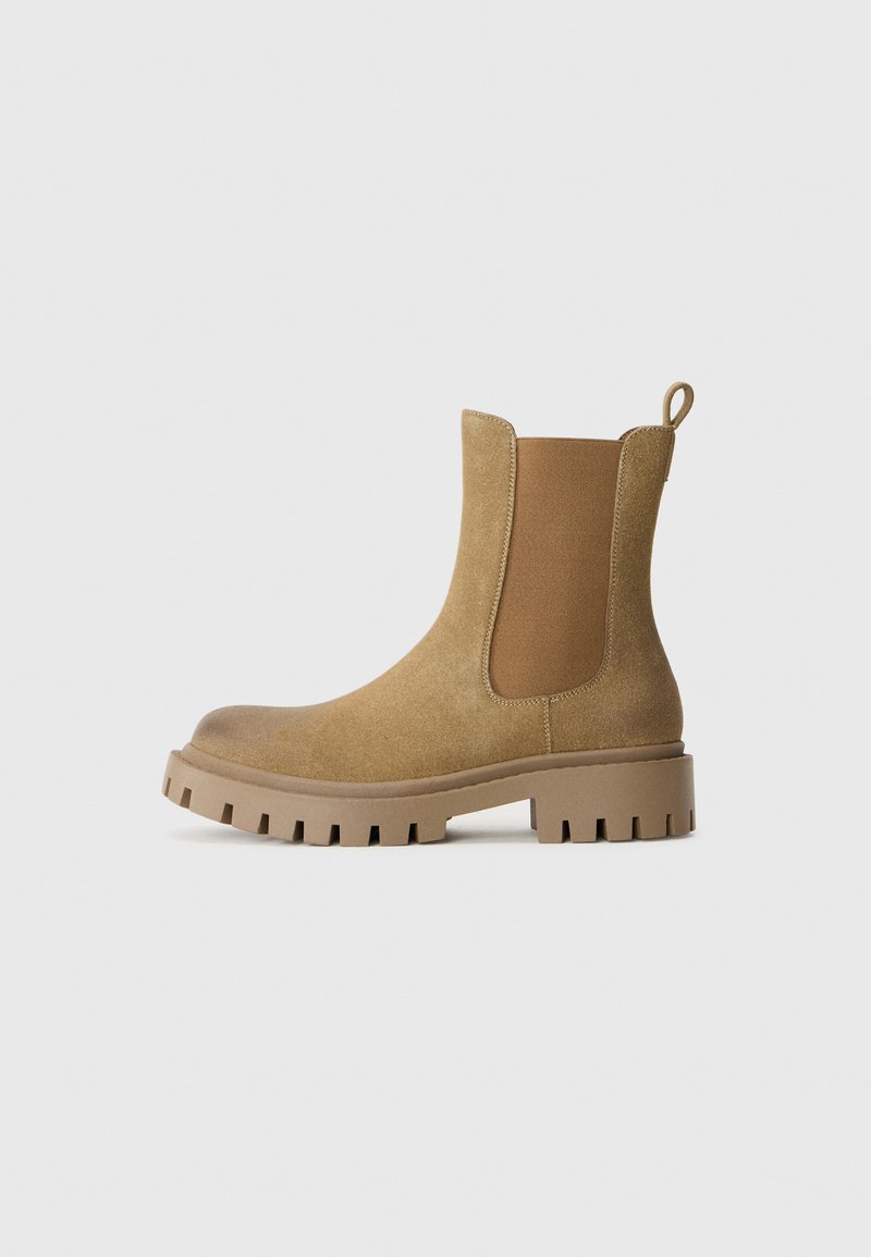 Botines tobilleros de gamuza en color beige, con suela de goma gruesa, paneles laterales elásticos y una lengüeta en el talón para facilitar el calzado.
