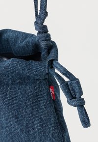 Σκούρο μπλε τζιν σακίδιο με κορδόνια και δεμένα λουράκια, με μια μικρή κόκκινη ετικέτα ραμμένη στη πλευρική ραφή, σε απλό λευκό φόντο.