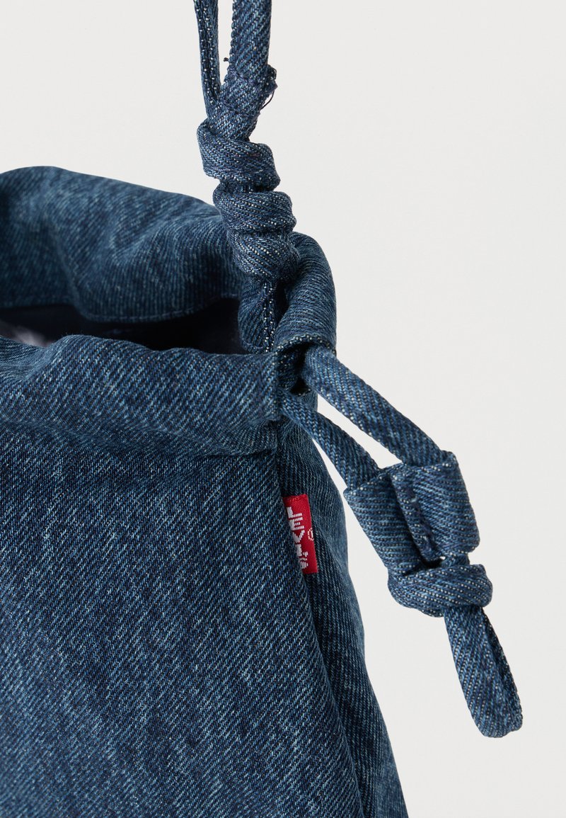 Borsa con coulisse in denim blu scuro con tiranti annodati e una piccola etichetta rossa cucita nella cucitura laterale su sfondo bianco semplice.