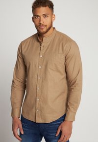 Camisa de manga larga de color marrón claro con cuello abotonado, bolsillo frontal y una tela texturizada. Combinada con pantalones vaqueros de mezclilla azules.