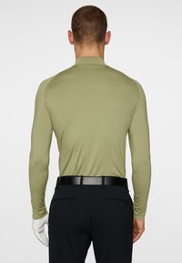 Långärmad, slim fit skjorta i olivgrön, tillverkad av ett mjukt och elastiskt tyg. Har en hög krage och bärs med mörka byxor och ett svart bälte.