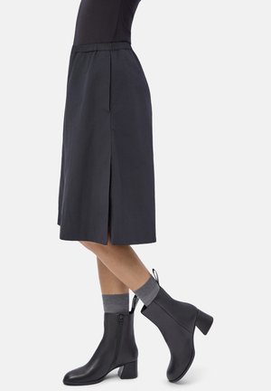 Person wearing a knee-length dark skirt with a side slit, gray socks, and black mid-heel ankle boots standing against a white background.