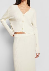 Elfenbensfärgad ribbad cardigan med V-ringning, kort längd och tre knappar. Matchas med en ribbad nederdel som visar en strukturerad stickad design.