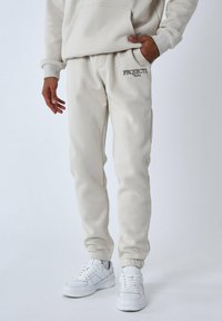 Pantalones de chándal beige hechos de una mezcla de algodón, con un logo en la parte delantera y puños elásticos. Combinados con zapatillas blancas.