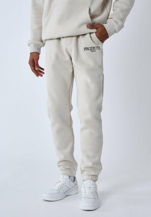 Panta felpa beige realizzati in tessuto misto cotone, con un logo sul davanti e polsini elastici. Indossati con sneaker bianche.