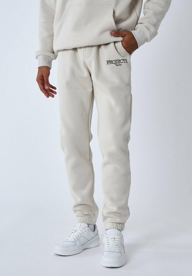 Pantalones de chándal beige hechos de una mezcla de algodón, con un logo en la parte delantera y puños elásticos. Combinados con zapatillas blancas.