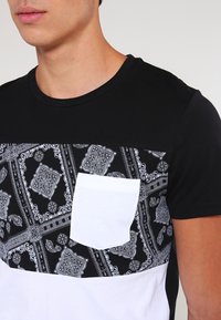Muž v tričku s černo-bílým vzorem a bílou náprsní kapsou, s krátkými rukávy, zobrazený od krku po střed trupu.