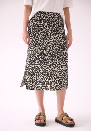 Personne portant une jupe midi beige et noire à imprimé léopard, un top blanc et des sandales compensées marron avec des boucles, debout sur un fond clair uni.