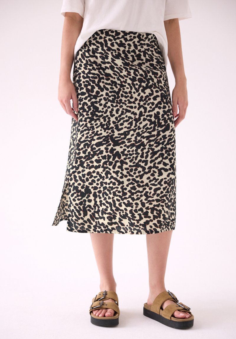 Personne portant une jupe midi beige et noire à imprimé léopard, un top blanc et des sandales compensées marron avec des boucles, debout sur un fond clair uni.