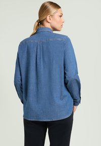 Koszula dżinsowa z kołnierzykiem, długimi rękawami i plisą z tyłu. Materiał w średnim odcieniu niebieskiego, o gładkiej teksturze, wykończony zaokrąglonym dołem.