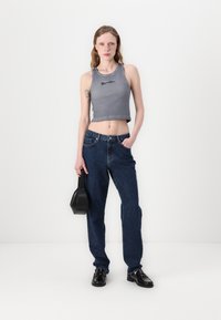 Grijze cropped tanktop met logo, donkerblauwe losse jeans, zwarte loafers, met een kleine zwarte handtas, alles tegen een neutrale achtergrond.