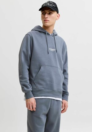 Jeune homme portant un sweat à capuche gris et un pantalon de survêtement assorti, une casquette noire, debout devant un fond clair uni, regardant sur le côté.