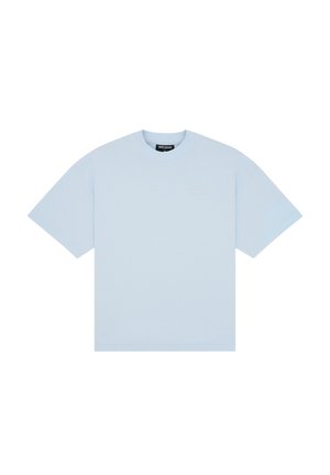Jeune femme portant un t-shirt bleu clair oversize, une mini-jupe plissée grise, des chaussettes blanches transparentes et des mocassins en cuir noirs, debout sur un fond uni.