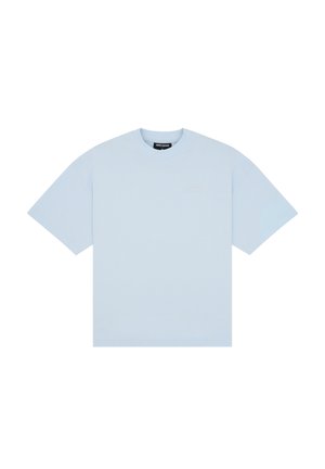 Młoda kobieta nosząca jasnoniebieski oversize T-shirt i szarą plisowaną mini spódniczkę, stojąca na jednolitym tle.