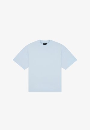 Młoda kobieta nosząca jasnoniebieski oversize T-shirt i szarą plisowaną mini spódniczkę, stojąca na jednolitym tle.